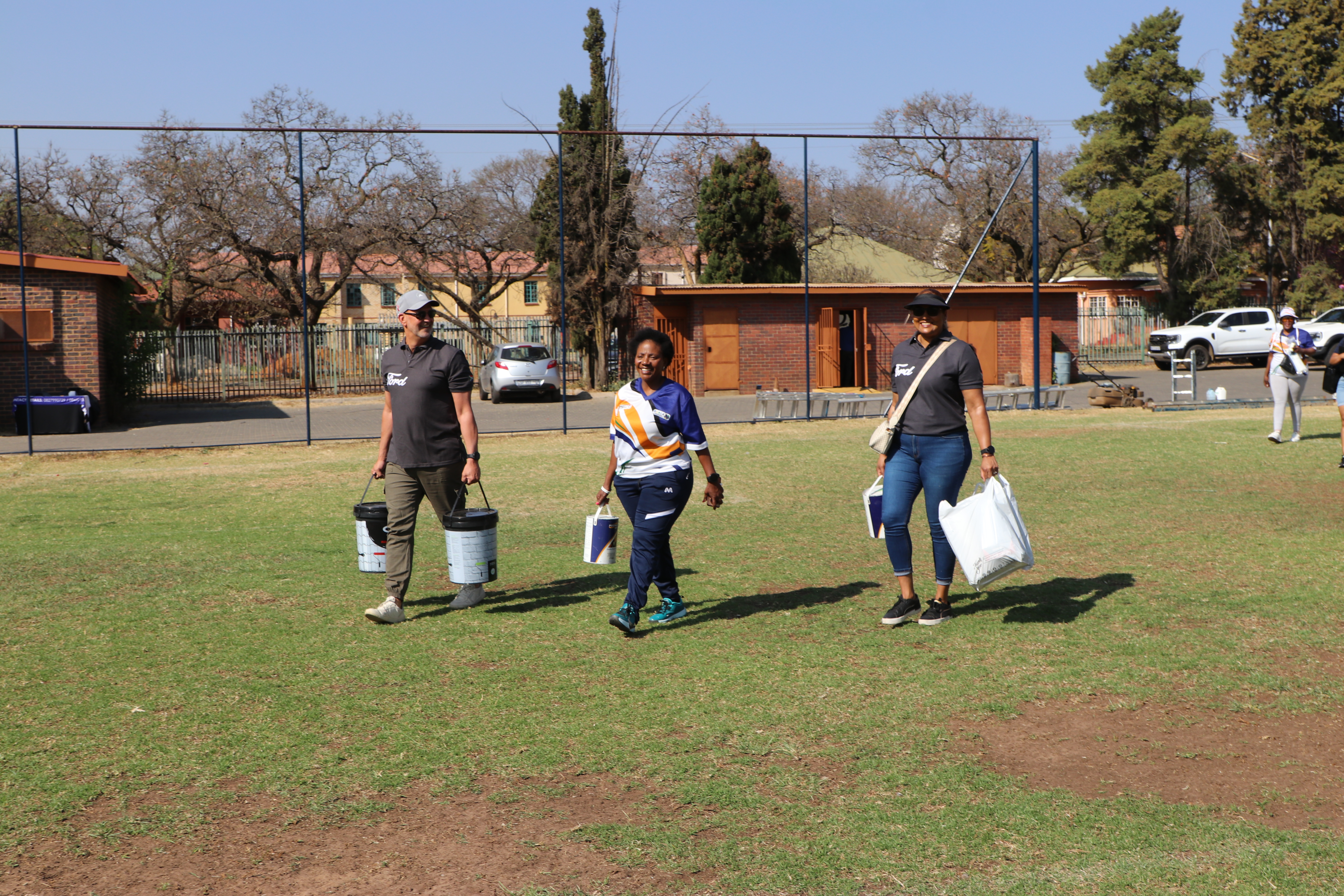 Three people carry items across a field with smiling faces.